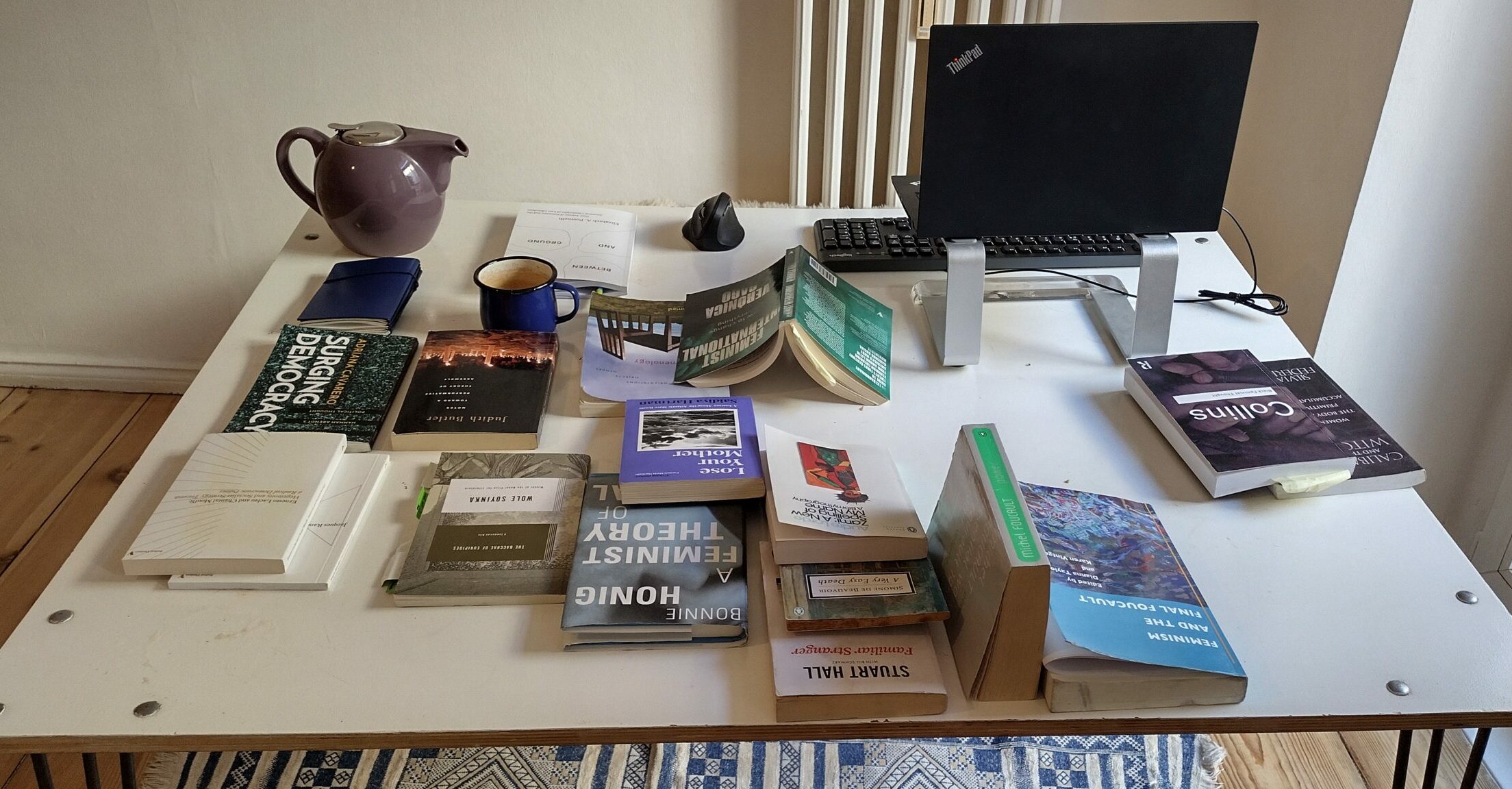 Das Bild zeigt den Schreibtisch von Liesbeth Schoonheim, auf dem sich mehrere aufgeschlagene Bücher, eine Tasse, eine Teekanne, Bildschirm und Tastatur befinden.