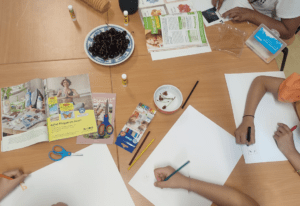 People drawing at a table with art supplies, cherries, and magazines.
