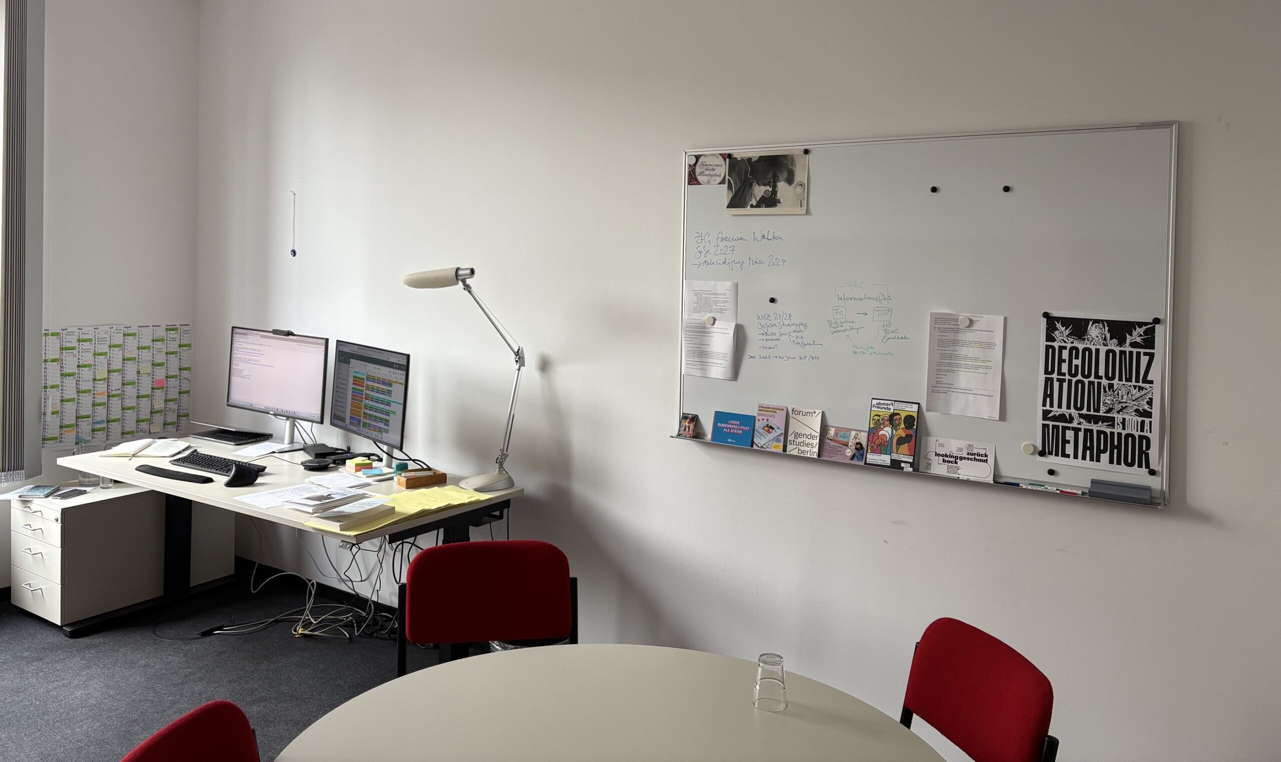 Büro mit Schreibtisch, zwei Monitoren, Tischlampe und diversen Arbeitsmaterialien. Rechts daneben Whiteboard mit handschriftlichen Notizen, angepinnten Dokumenten und einem Poster mit der Aufschrift „DECOLONIZATION IS NOT A METAPHOR“. Im Vordergrund runder Tisch mit drei roten Stühlen. Wandkalender an der linken Seite. Kabel unter dem Schreibtisch sichtbar. Tageslichtbeleuchtung.